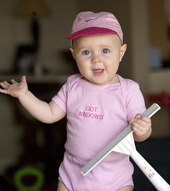 A baby wearing a pink outfit and cap holds a window squeegee in one hand and has the other hand raised, promoting her Dad's window cleaning company.